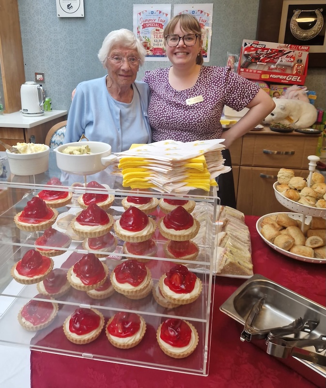 Elderly woman and young woman smile behind a counter stocked with cakes